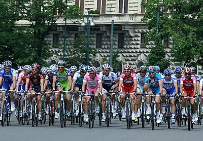 Cyclists from many teams riding in the streets. A building, streetlamps and trees are shown in the background.