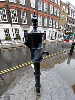 An old-fashioned water pump, painted black, with its handle removed, standing on a stone pedestal two steps above the sidewalk.