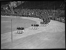 Black and white photo of the seating area of a single-tiered stadium, surrounding a running track with athletes parading in a line