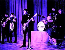 A tinted photograph of five members of the D-Men performing with guitars, drums, and keyboards