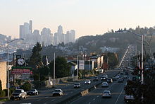 A divided highway seen alongside several buildings, with a city skyline in the background behind a hill.