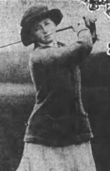 A white woman swinging a golf club; she is wearing a brimmed hat, dark sweater, and light-colored skirt