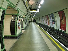 View along a platform in a circular tunnel. Tracks run on the right side with the walls covered with cream and green tiles. An illuminated 'way out' sign overhead indicates the exit.