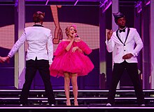 Photograph of Minogue wearing in a pink bra and red panties holding a mic with two male dancers to her sides
