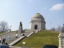 Bâtiment circulaire blanc surmonté d'un dôme au sommet d'une butte avec un escalier d'accès où se trouve une statue noire.