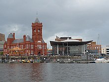 Red brick Victorian style building with clock tower and the Senedd building, with water in foreground