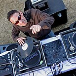 A man wearing sunglasses, standing in front of a turntable and pointing upwards