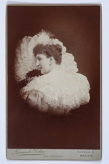 A white woman with curled hair, wearing a lace-trimmed white dress, posed in front of a feathery or ruffled fan