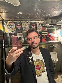 A photo of the author Anthony Oliveira, a smiling white man with black hair and a light beard. He is wearing a black jacket with several pins and a white shirt with a sacred heart on it.