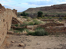 A partly overcast sky and subdued sunlight over a roughly six-foot-tall wall of dusky tan sandstone bricks which vary somewhat in size. The wall runs diagonally from the immediate foreground at left towards the right, running perhaps several dozen feet to the near middle distance. A few feet to the right, in the middle foreground, a low ring of similar blocks delimits a circular pit sunk into the ground. The remains of several other ruinous low walls, perhaps one to three high at most, are arrayed in parallel; they align left to right from the high diagonal wall. Perhaps a mile distant to the center and right, a canyon wall slopes gradually level to meet the valley floor on which the walls sit.