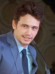 A head shot of James Franco, a caucasian male in his mid-20s with dark hair, looking slightly away from the camera and smiles. He wears a blue suit with a light blue shirt and dark blue tie, and speaks into a mic.