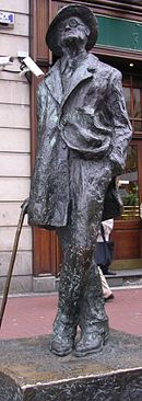 Bronze statue of Joyce standing in a coat and broadbrimmed hat: His head is cocked looking up, his left leg is crossed over his right, his right hand holds a cane, and his left is in his pants pocket, with the left part of his coat tucked back.