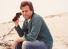 Photograph of Mark Lesser sitting on beach dunes, holding binoculars in one hand and smiling at the camera.