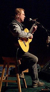 A fair-haired man sitting on a stool playing a guitar