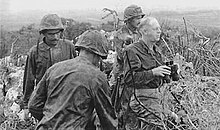 A black-and-white image of Merritt Edson and three other Marines standing in bushes. Edson is holding some binoculars and is looking out over the battlefield.