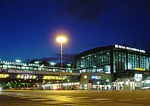 The exterior of a railway station at night. A rectangular building on the right has 2 of its sides visible, both of their walls windowed. A shorter connecting structure on the left is suspended over a street and lined with advertisements.