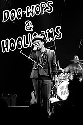 A black and white picture of Bruno Mars fully dressed in a suit and a hat singing for a crowd.