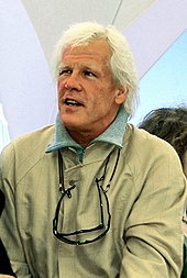 A man with white hair, wearing a tan jacket and two pairs of glasses on strings around his neck, looks off-camera.