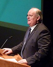 Man in his 50s wearing glasses and a suit standing at a lectern