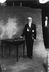 Photo d’un homme chauve et à la barbe blanche se faisant photographier debout, en tenue officielle de chef de l’État, la main droite sur deux livres posés sur une table