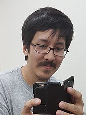 Ronald Watkins pictured from the shoulders up, wearing a gray t-shirt and glasses. He is holding and looking at a cell phone in a black folding case.