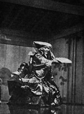 Acteur en tenue traditionnelle, masque féroce, bras en avant tenant un éventail. Noir et blanc.