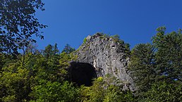 Photographie dans une forêt de l'entrée d'une grotte avec une ouverture assez large sur une paroi rocheuse.
