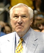 An older Caucasian man with white hair is standing up on a basketball court. He is wearing navy blue dress pants, a baby blue suit jacket with a white cross-hatching pattern, and a bright yellow tie.