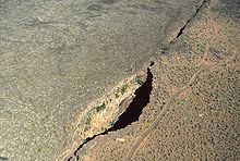 A large, deep hole in the ground along a visible fault line viewed from the air. The bottom cannot be seen.