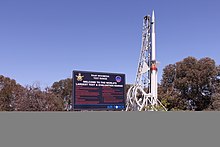 A white Long Tom rocket stands vertically on its launch gantry alongside a sign welcoming visitors to the Woomera Test Range