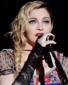 Madonna singing to a microphone wearing a colorful blouse with see-through black sleeves and gloves.