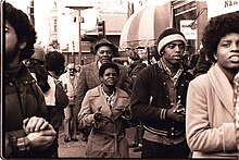 A smiling group of mostly African-American standing in the street in front of a building clapping their hands.