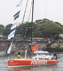 Voilier orange et blanc descendant le chenal. Le navigateur, debout à l'avant, salue la foule.