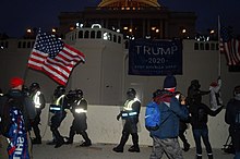 A photo taken near dusk of the Capitol building. A large Trump flag is hung on a railing. Protestors are still remain, and a small line of police officers yield shields.