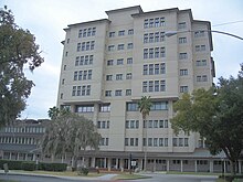 a ten-story building with sable palms, a Spanish moss-drapped oak tree, hedges, and a court yard