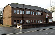 Three-quarter view of a two-storey brown brick building with a shallow, grey, partly hipped roof. The two storeys are separated by a thin band of projecting bricks. Each floor has three sets of four rectangular windows. To the right, a projecting section includes a round-headed entrance door, the words "BOWLS ENGLAND" on a white background, and a red and blue logo consisting of a heraldic lion and a crown.