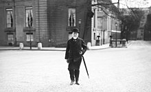 Photo en noir et blanc d'un homme à barbe blanche marchant dans une cour, avec un chapeau sur la tête et un parapluie à la main