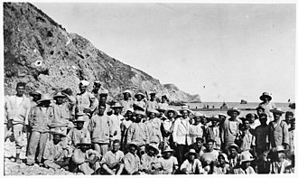 a group of Italian sailors standing near the shore with naval ships in the background