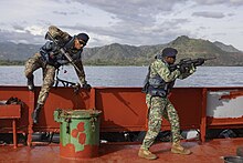 Colour photo of two men wearing military uniforms and carrying guns on the deck of a ship