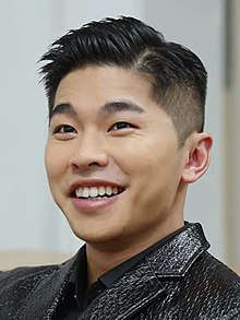 Close up of Yau dressed in a black jacket and smiling while seated on a sofa.