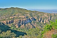 Landscape of dry rugged hills with areas of exposed rock, steep cliffs, and low vegetation, below the sky