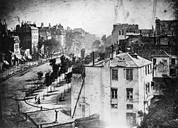 ダゲールによる写真「タンプル大通り（Boulevard du Temple）」。1838年から1839年に撮影。10分以上の露光により、道路を行き交う人馬は全く写っていないが、唯一画面左下に片足を何かに乗せた人物が写っており、人類史上初めて写真に写った人物とされている。