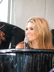 A blonde woman smiles as she looks to her right while sitting behind a piano