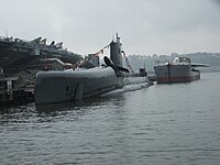 A submarine tied up at a wharf with an aircraft carrier alongside and a barge containing Concorde behind