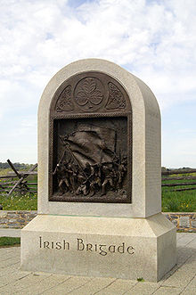 A tall stone monument with a rounded top and a bronze inset sculpture. The base reads, Irish Brigade.