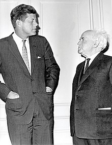 A candid portrait of Kennedy and Ben-Gurion standing facing each other, dressed formally, Kennedy with his hands in his suit jacket pockets.