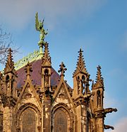 Photographie en couleurs de la statue et d'une partie du faîtage depuis le sol entourés de tabernacles et de gargouilles.