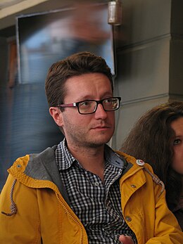 Photograohie en couleurs d'un homme en buste, avec lunettes et imperméable jaune.