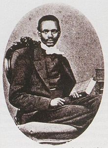 Black and white image of a seated black man with close cropped dark hair facing the camera, wearing a dark suit with a light coloured cravatte and holding a book in his left hand.