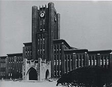 Photo noir et blanc d'un bâtiment en pierre étendu sur toute la largeur du cliché, avec une tour centrale exhibant une horloge sur chaque façade visible, sur fond de ciel clair.